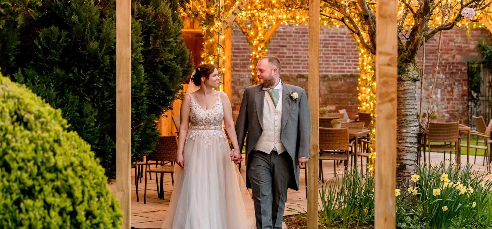 Bride and Groom walking hand in hand away from the pergola lit up with orange lights