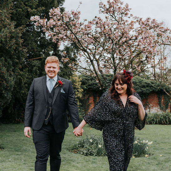 Bride in a dark coloured dress walking away from a tree in blossom hand in hand with the grrom